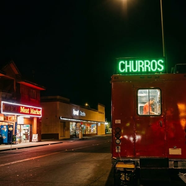 A red churro food truck with a green neon sign at night, parked on a street with people nearby.